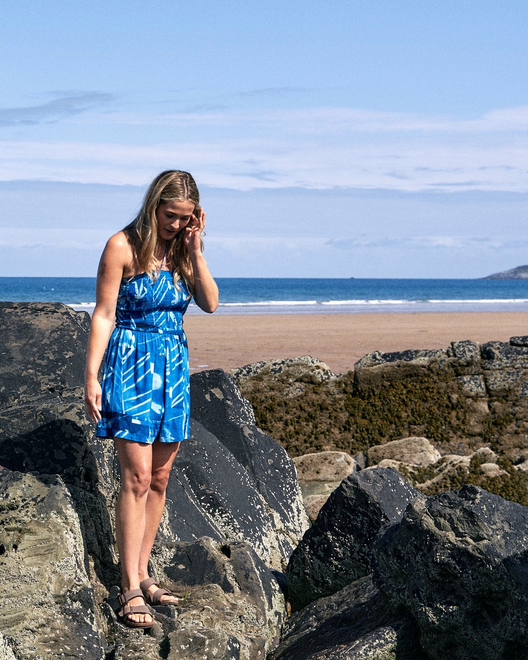 A woman in a blue dress stands on rocks at a beach with the ocean and distant hills in the background, her Hesta - Womens Playsuit - Blue by Saltrock gently swaying in the breeze.