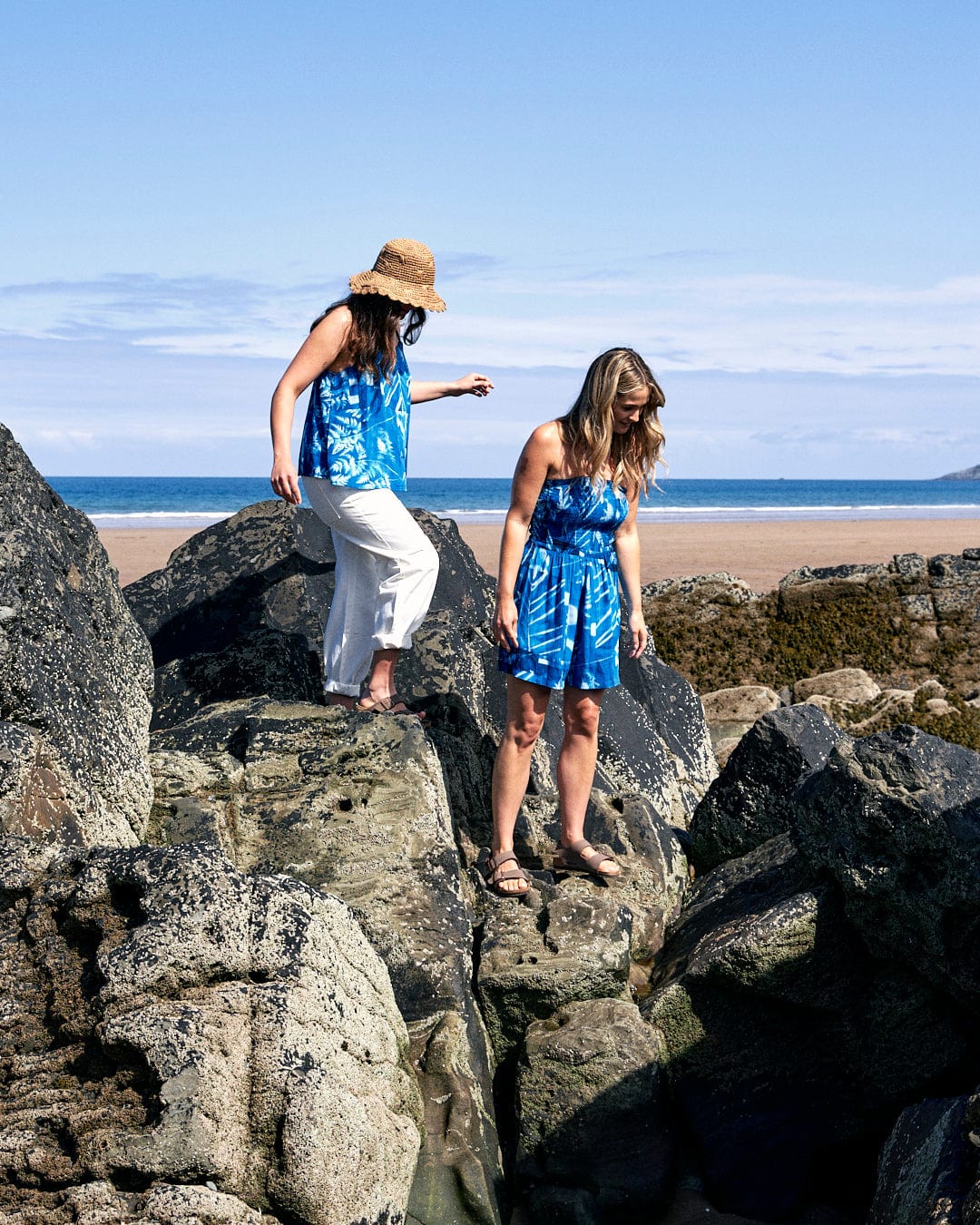 Two women wearing blue outfits, including a crinkle viscose dress with an abstract floral print and culotte shape shorts, stand on large rocks with the beach and ocean in the background. One wears a hat and white pants, while the other wears a Saltrock Hesta - Womens Playsuit - Blue.