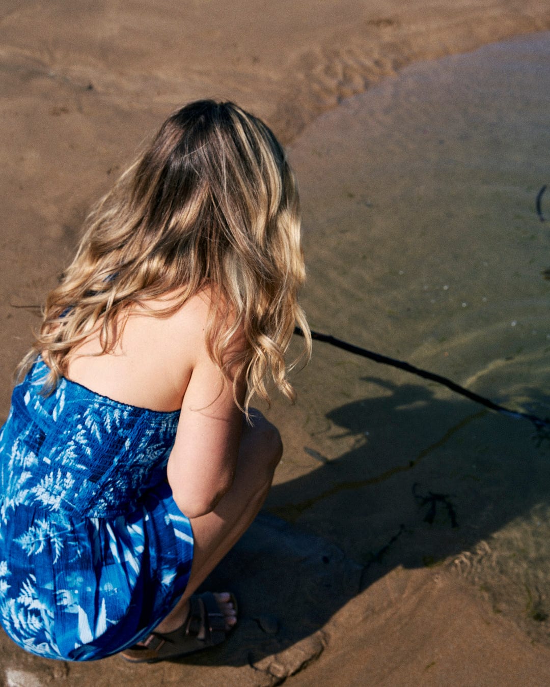 A woman with long blonde hair, wearing a Saltrock Hesta - Womens Playsuit - Blue with an abstract floral print, crouches on the sand at the edge of water, holding a stick.