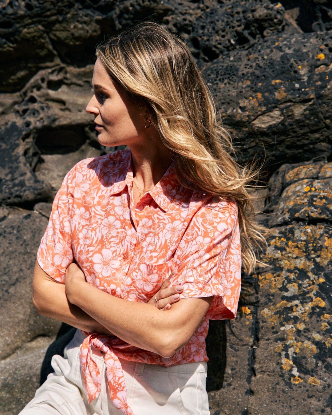 A woman with long hair in a Saltrock Hibiscus - Womens Tie Front Shirt - Orange and white pants stands with crossed arms, looking to the side in front of a rocky background.