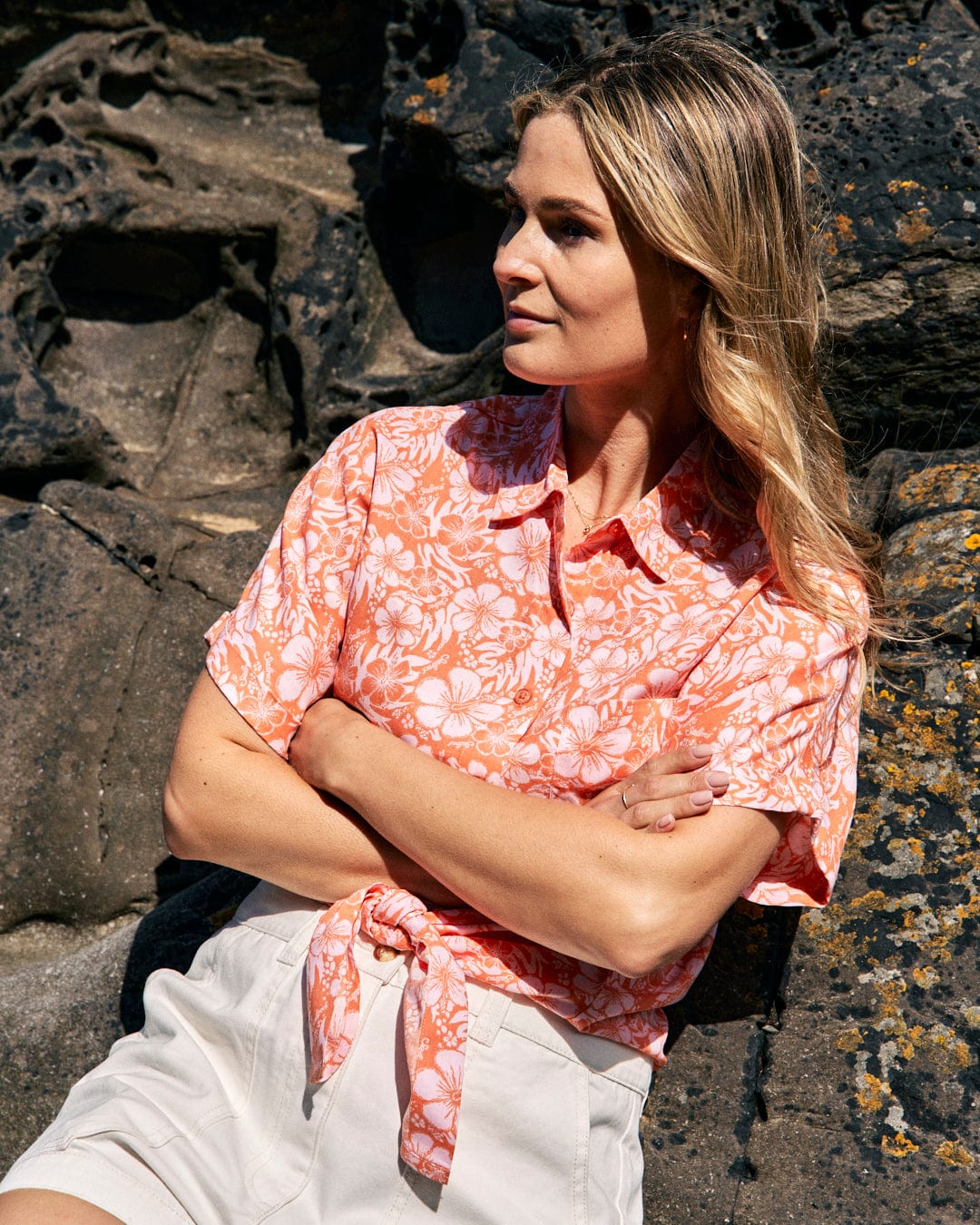 A woman wearing a Saltrock Hibiscus - Womens Tie Front Shirt - Orange and light-colored shorts stands with arms crossed in front of a rocky backdrop.