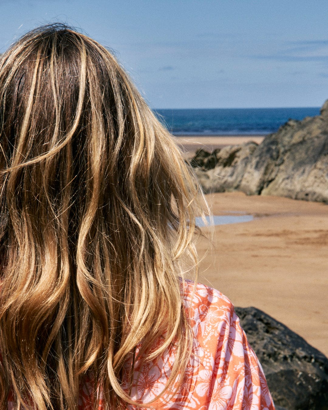 A person with long blonde hair, wearing a Saltrock Hibiscus - Womens Tie Front Shirt - Orange in lightweight material, stands facing a beach with rocky cliffs in the distance.