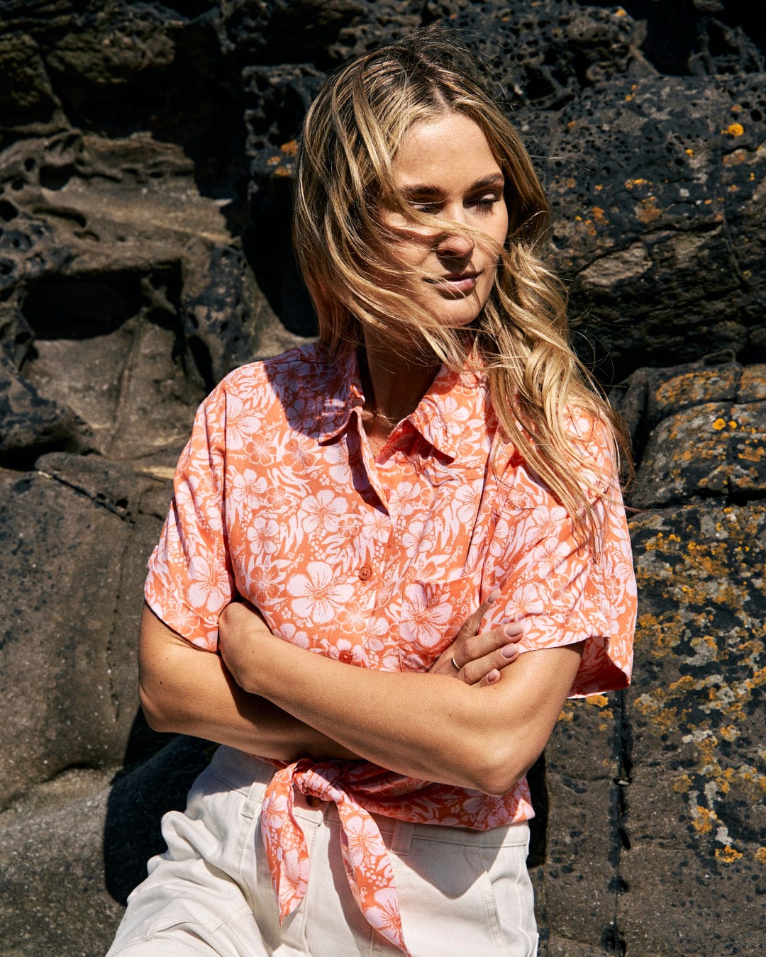 A woman with long blonde hair stands against a rocky background, wearing a Saltrock Hibiscus - Womens Tie Front Shirt - Orange, looking to her side.