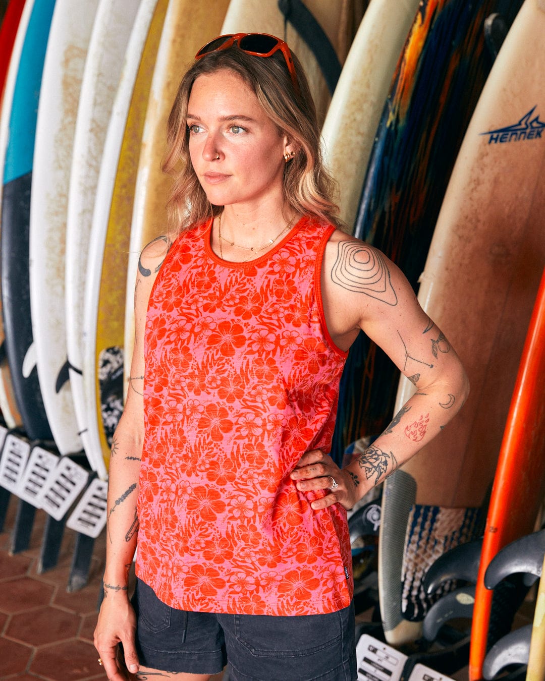 A woman stands in front of a rack of surfboards, wearing a Saltrock Hibiscus - Womens Vest - Red and black shorts, one hand on her hip. She has sunglasses perched on her head and various tattoos on her arms.