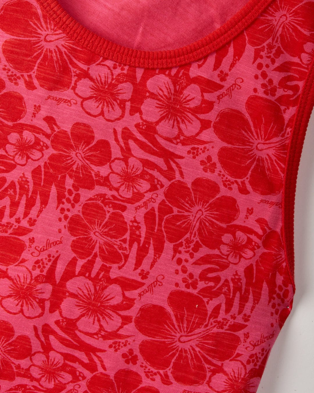 Close-up of a red and pink floral-patterned fabric featuring a vibrant hibiscus print, with the word "Saltrock" repeated throughout. The 100% cotton material is machine washable for easy care. This is the "Hibiscus - Women's Vest - Red" by Saltrock.