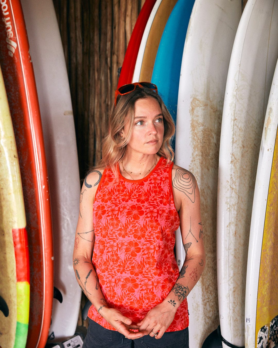 A woman with tattoos, donning a Saltrock Hibiscus - Womens Vest - Red and sunglasses on her head, stands in front of a row of surfboards.