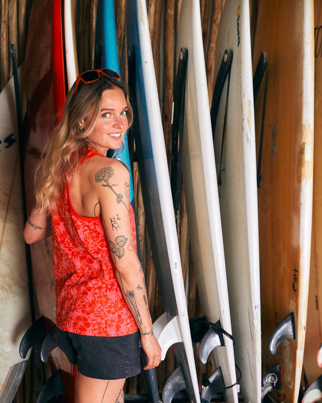 A person with tattoos, wearing a Saltrock Hibiscus - Womens Vest - Red and hibiscus print shorts made of 100% cotton soft peached fabric, stands in front of a rack of surfboards and smiles while looking back at the camera.