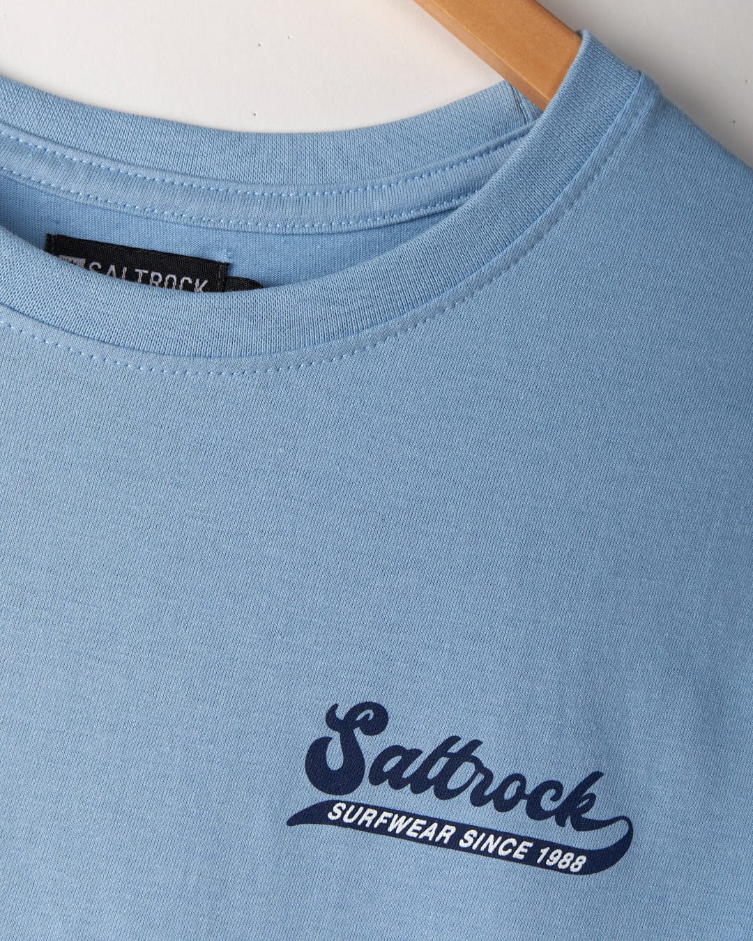 Close-up of a light blue Home Run - Mens T-Shirt - Blue on a hanger, crafted from 100% cotton with the brand's logo and the text "Saltrock Surfwear Since 1988" in dark blue on the chest.