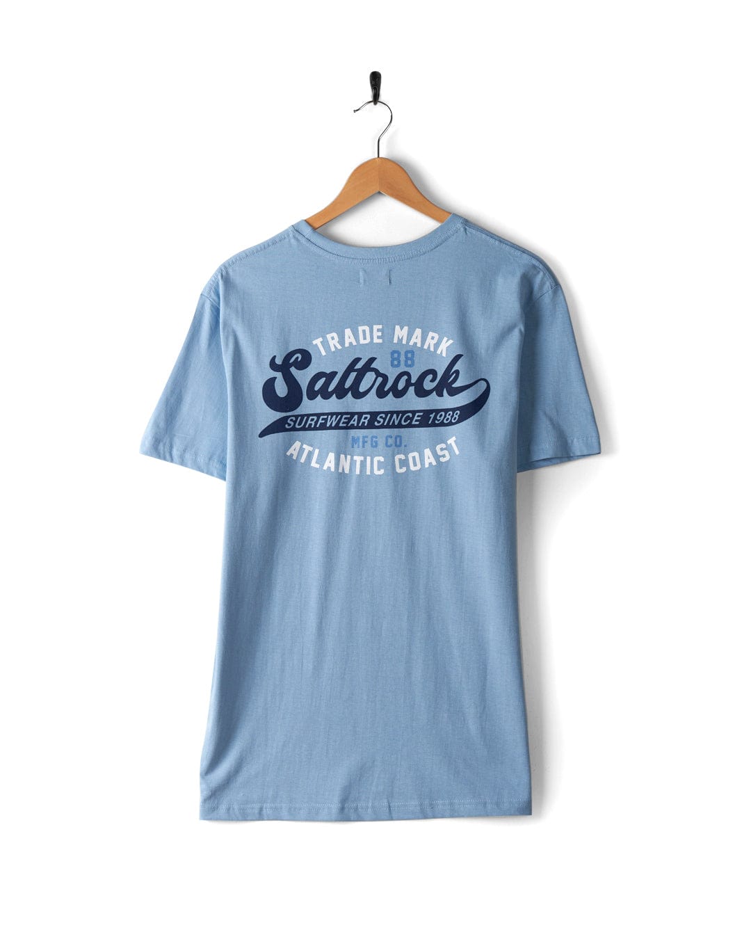 A light blue Home Run - Mens T-Shirt - Blue, made from 100% cotton, featuring a “Saltrock” logo and “Surewear Since 1968 MFG Co. Atlantic Coast” text printed on the back, hangs gracefully on a wooden hanger.