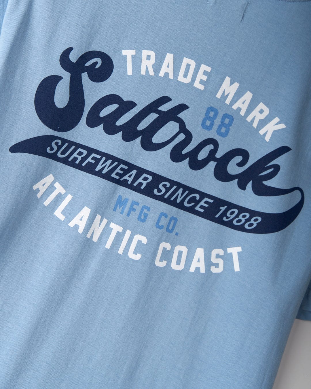 Close-up of a light blue t-shirt with the text "Saltrock Surfwear Since 1988. Trademark MFG Co. Atlantic Coast." written in a curved format, showcasing classic Saltrock branding. This is the Home Run - Mens T-Shirt - Blue from Saltrock.