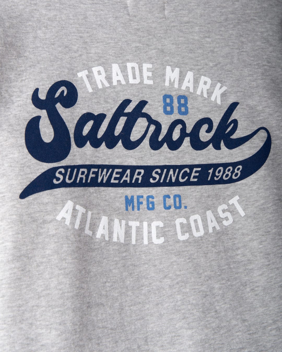 Close-up of a grey color **Home Run - Mens Zip Hoodie** with the text "Saltrock Surfwear Since 1988 Atlantic Coast" and "Trade Mark MFG CO" printed in blue and white.