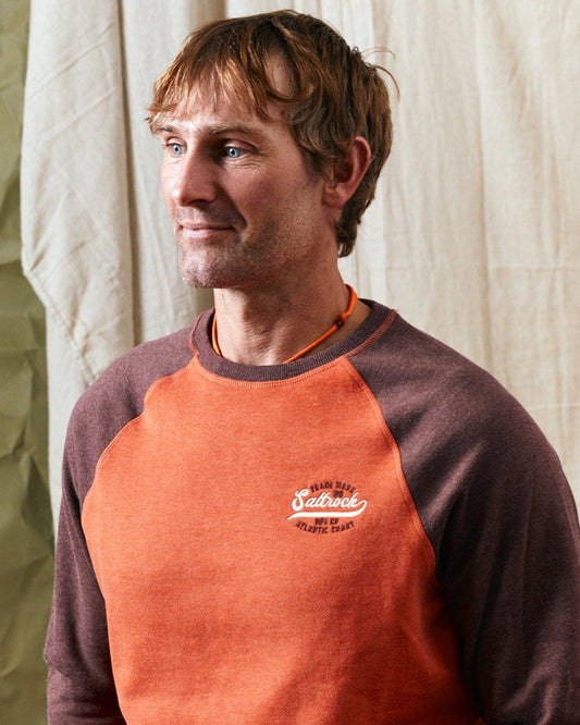 A man with short brown hair and light stubble wears the Home Run Raglan - Mens Sweat - Orange by Saltrock, featuring the brand's name embroidered on the chest. He stands in front of a light fabric backdrop.