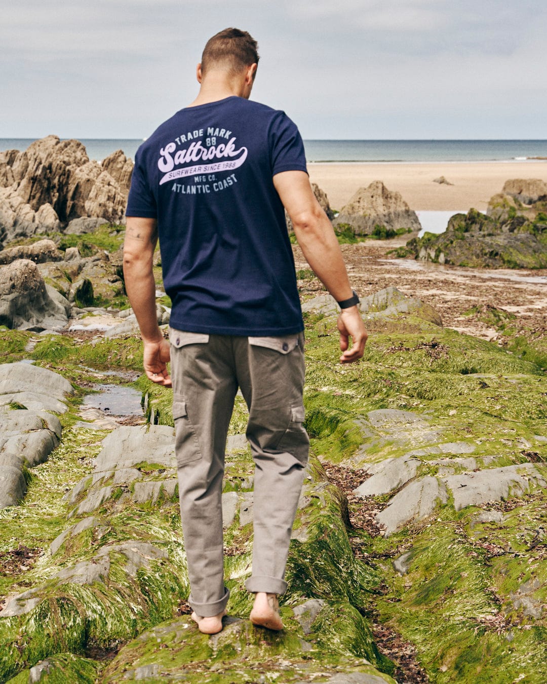 A man in a Home Run dark blue, 100% cotton, Saltrock-branded t-shirt and gray pants walks barefoot on mossy rocks by the beach with the ocean in the background.