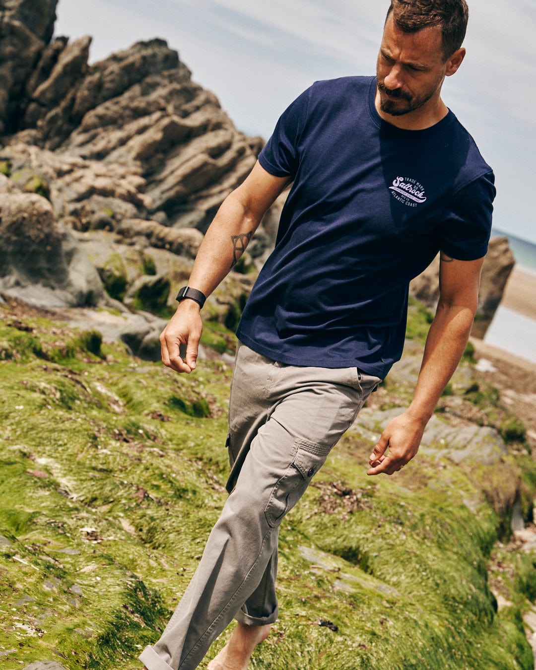 A man in a dark blue Home Run short sleeve t-shirt by Saltrock and beige pants walks barefoot on a rocky, moss-covered beach.