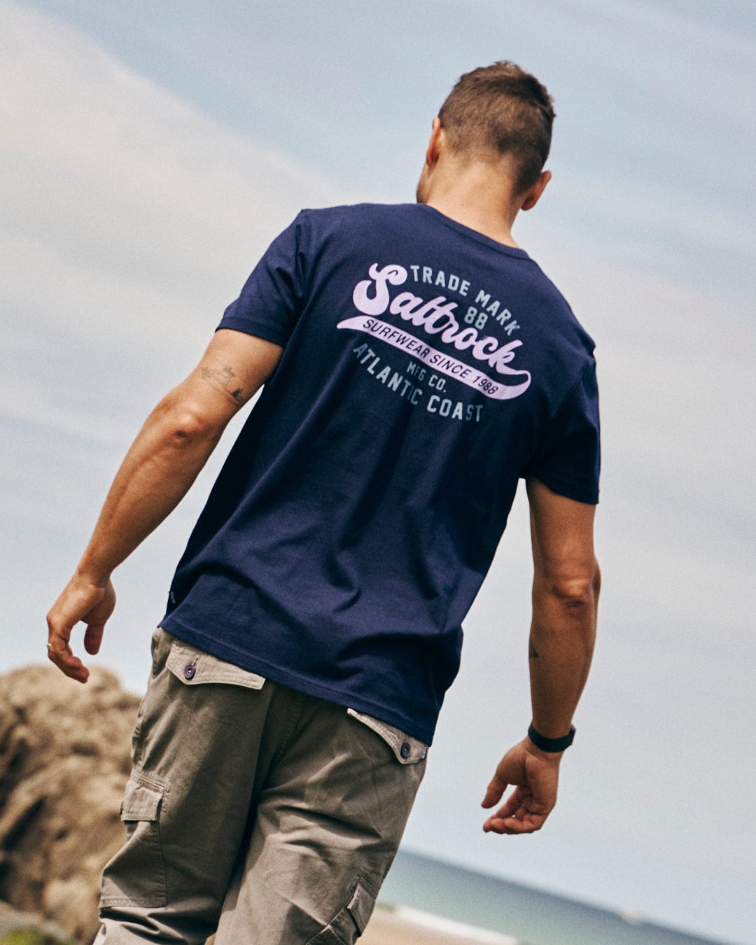 A person wearing a "Saltrock" Home Run men's short sleeve t-shirt in dark blue, made from 100% cotton, and khaki cargo shorts walks along a sandy beach.