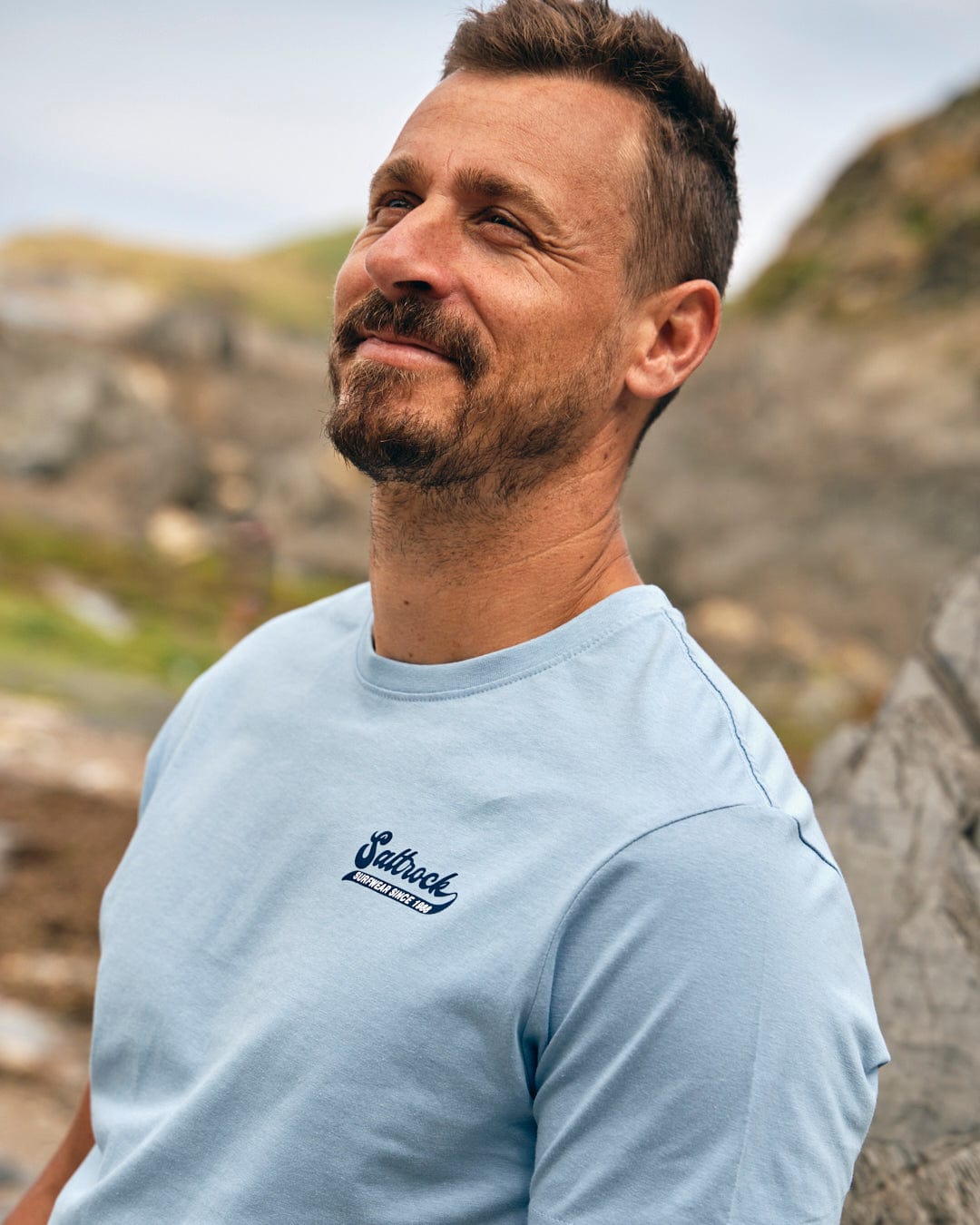 A man with short hair and a beard smiles while wearing the Home Run - Mens T-Shirt - Blue made by Saltrock, outdoors with rocks and greenery in the background.