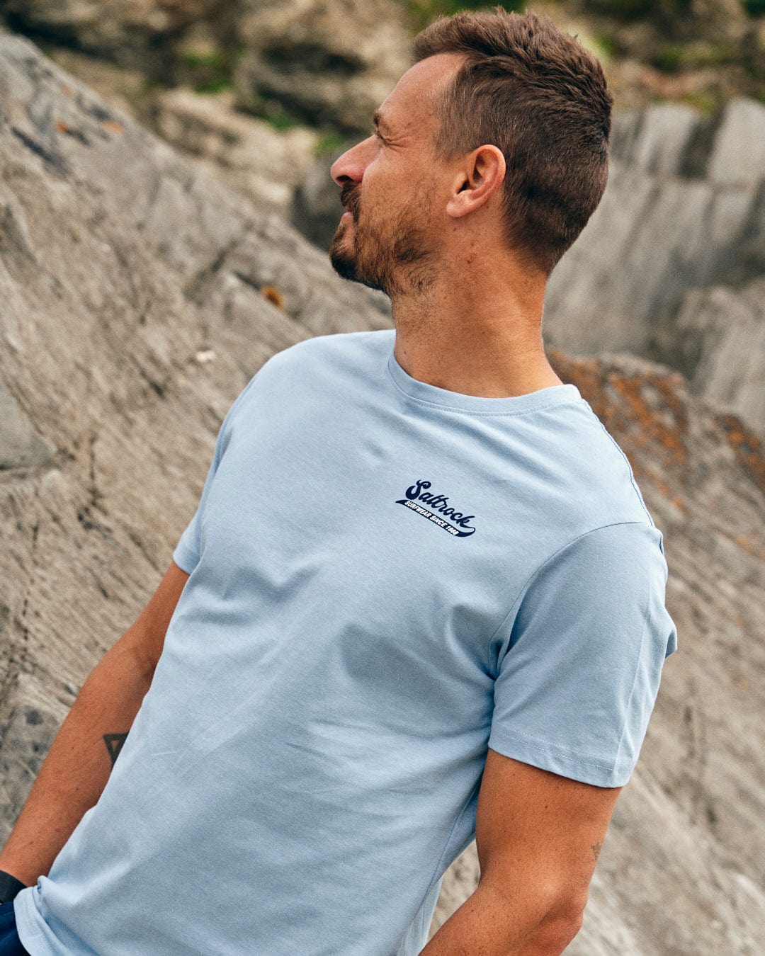 A man in a light blue, 100% cotton Saltrock Home Run - Mens T-Shirt - Blue stands outdoors with a rocky background, looking to his right.