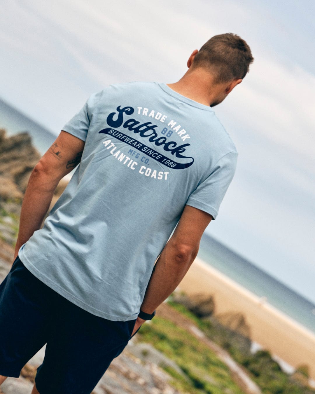 A man is standing on a rocky beach, gazing at the ocean. He is wearing a Home Run - Mens T-Shirt - Blue with Saltrock branding on the back and dark shorts.