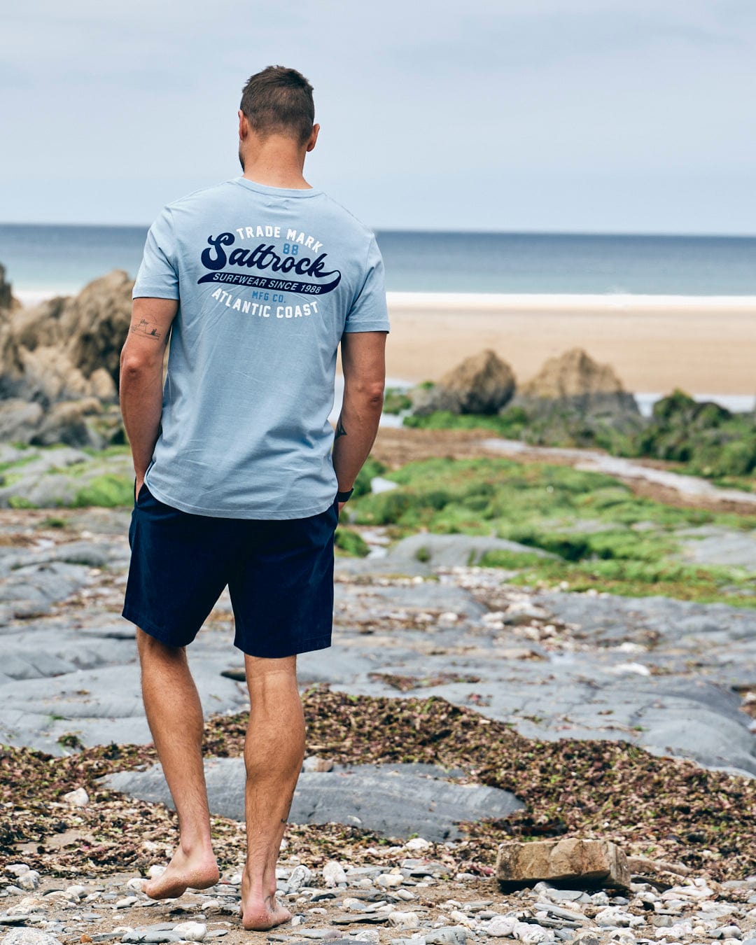 A man wearing a light blue, 100% cotton t-shirt and dark shorts stands on a rocky beach facing the ocean. The back of his Saltrock Home Run - Mens T-Shirt - Blue reads "Saltrock Atlantic Coast.