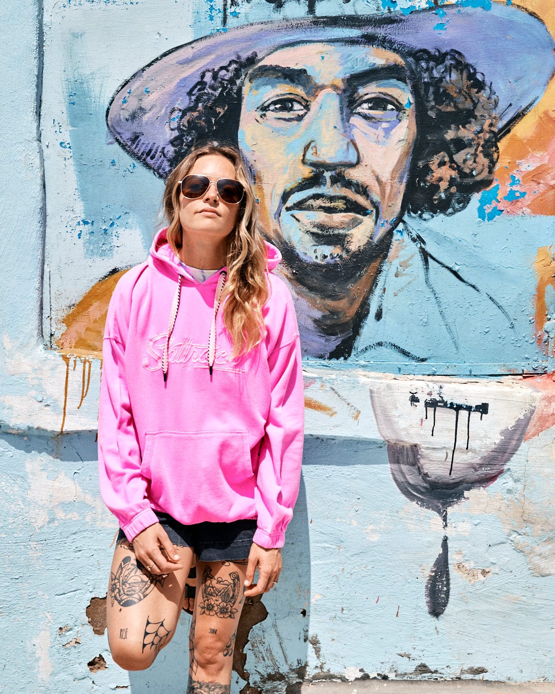 Person in a bright pink Instow - Womens Pop Hoodie - Pink and sunglasses stands in front of a mural painted on a wall, featuring a man's face wearing a hat. The person has tattoos on their legs and is sporting Saltrock branded short black shorts with an elasticated waist.