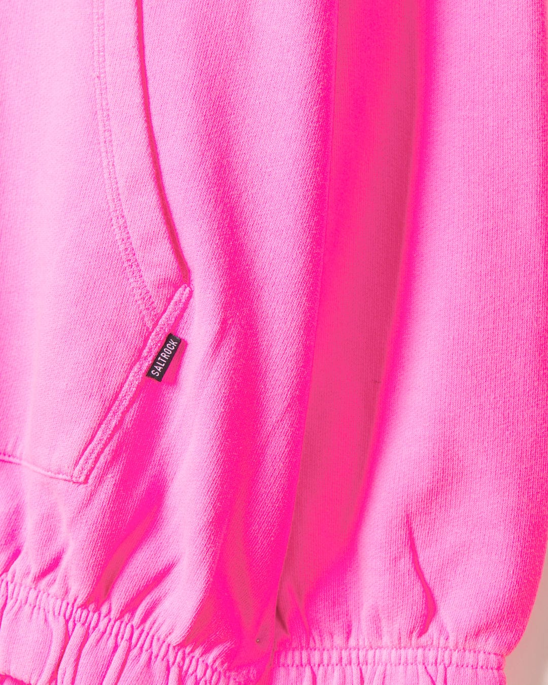 Close-up of a bright pink sweatshirt showcasing a pocket and a small black tag with text sewn into the seam near the bottom hem, made from 100% cotton. The product is the Instow - Womens Pop Hoodie - Pink by Saltrock.