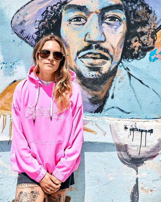 A woman wearing sunglasses and a bright pink **Saltrock Instow - Womens Pop Hoodie - Pink** with an elasticated waist stands in front of a wall mural depicting a man's face.