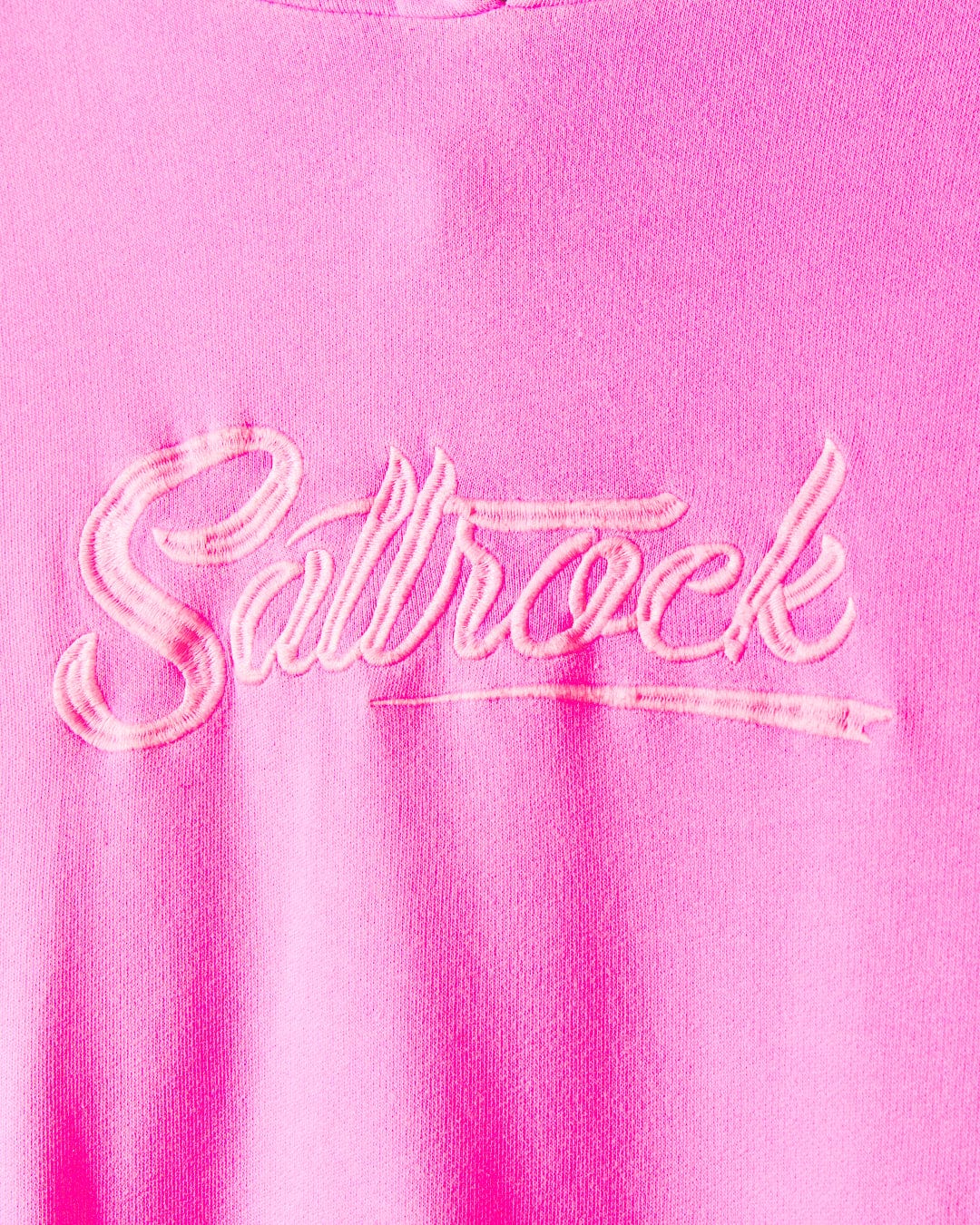 Close-up of a pink garment featuring "Saltrock" embroidered in stylized cursive stitching on the 100% cotton fabric, specifically the Instow - Womens Pop Hoodie - Pink.