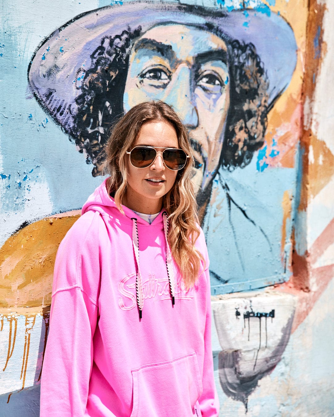 A person wearing a Saltrock branded Instow - Womens Pop Hoodie - Pink and sunglasses stands in front of a colorful mural featuring a person with curly hair and a hat.