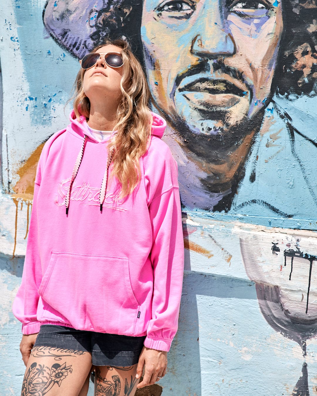 A person in an Instow - Womens Pop Hoodie - Pink and sunglasses, featuring an elasticated waist from Saltrock, stands in front of a mural featuring a man's face.