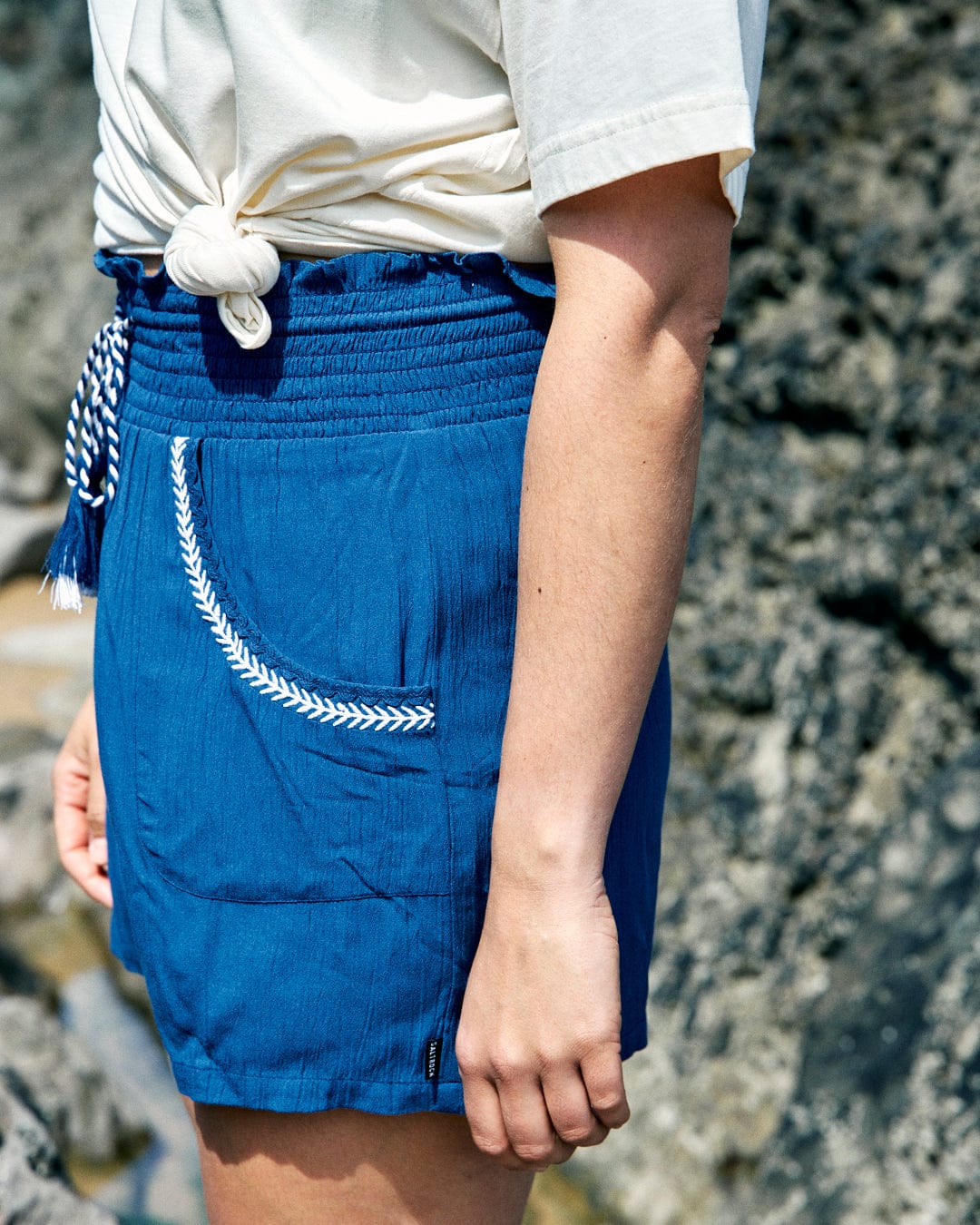 Person wearing a white T-shirt and Saltrock Jonie - Womens Shorts - Blue with an embroidered edging on the patterned pocket, standing outdoors near rocky terrain.