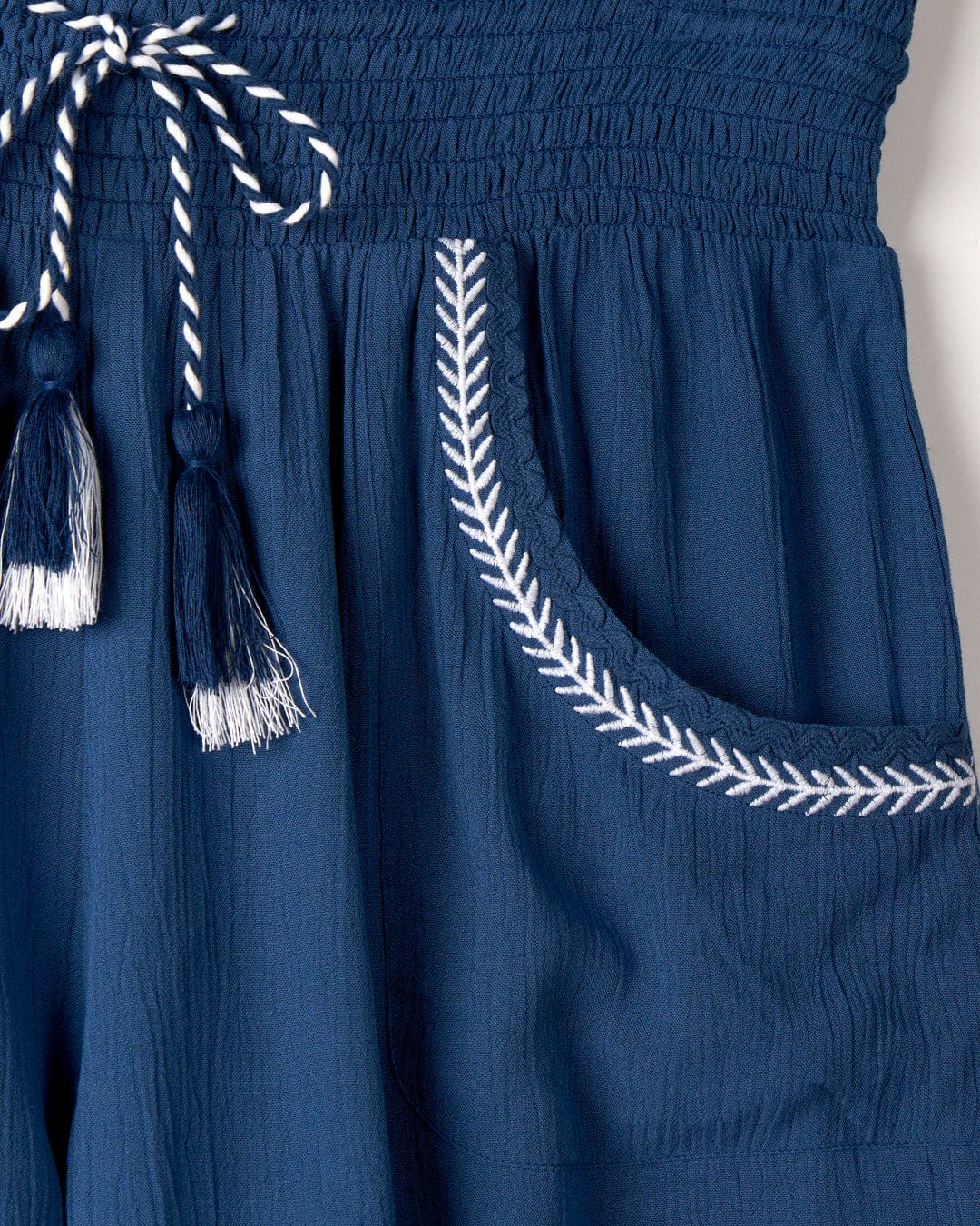Close-up of a blue fabric garment with a white embroidered pocket and tassel details on a drawstring. The lightweight material boasts a textured appearance, complemented by delicate embroidered edging. This is the Jonie - Womens Shorts - Blue by Saltrock.