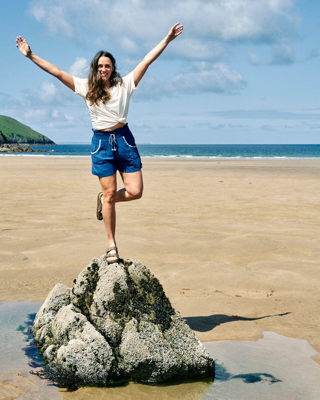 A woman stands on one leg atop a rock on a sandy beach with the ocean and a green hill in the background. She has her arms raised, smiles at the camera, and wears Saltrock's Jonie - Womens Shorts - Blue.