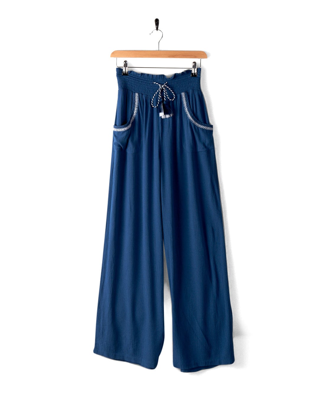 Jonie - Womens Trouser - Blue by Saltrock in a lightweight crinkle material, featuring an elastic high waistband with a decorative tie at the front and two front pockets, hanging on a wooden hanger.