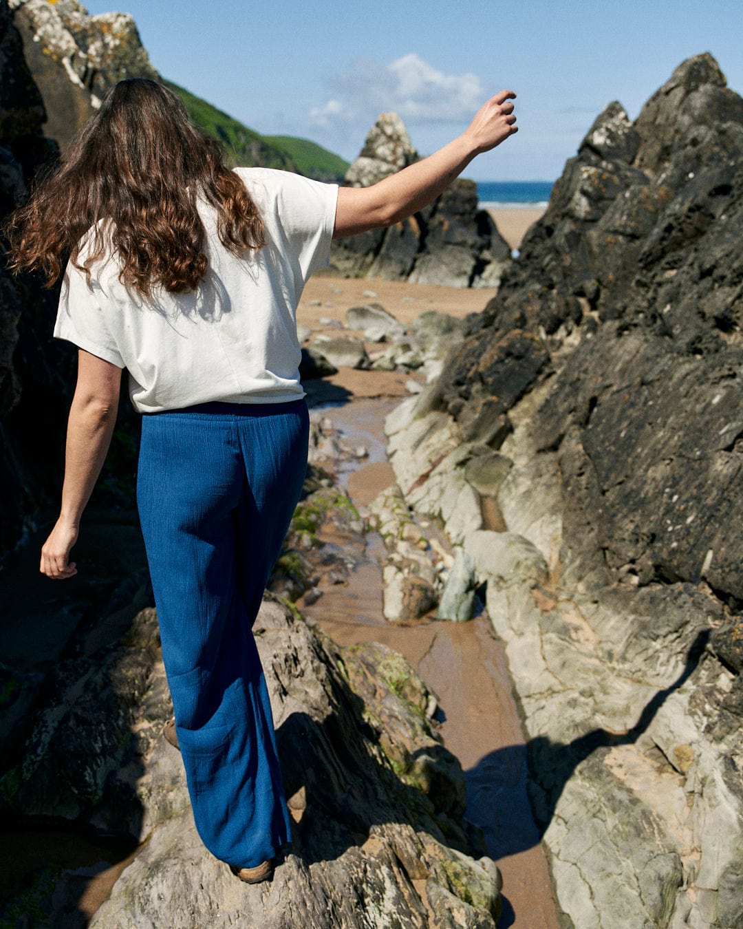 A person with long hair in a white shirt and Jonie - Womens Trouser - Blue from Saltrock walks over rocks near a beach with green hills and a blue sky in the background.
