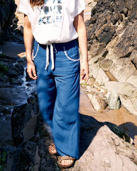 Person in a white crinkle viscose t-shirt and Saltrock Jonie - Womens Trouser - Blue with an elasticated waistband standing on rocky terrain near a beach.