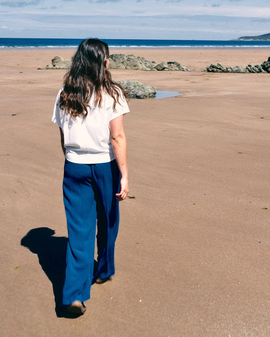 A person with long hair walks on a sandy beach, facing away, wearing a white top and high-waist Saltrock Jonie - Womens Trouser - Blue made of lightweight material. Rocks and the ocean are visible in the background.