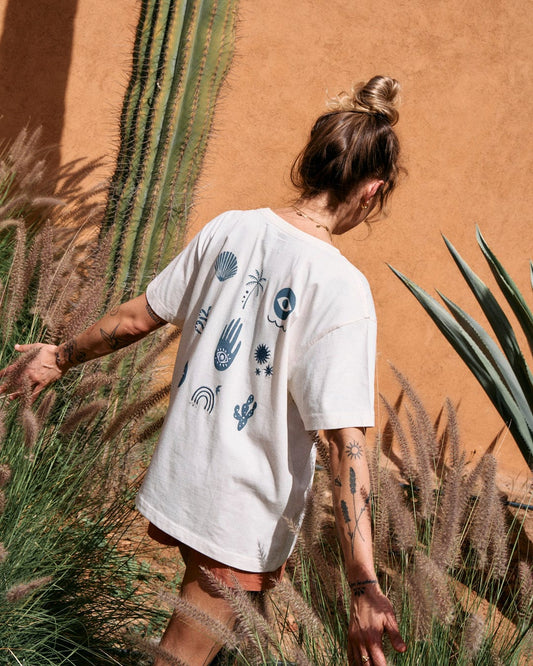 A person with a bun hairstyle, wearing a Saltrock Journey - Recycled Womens Short Sleeve T-Shirt - Cream adorned with various symbols, is walking through tall grass and plants next to a terracotta-colored wall.