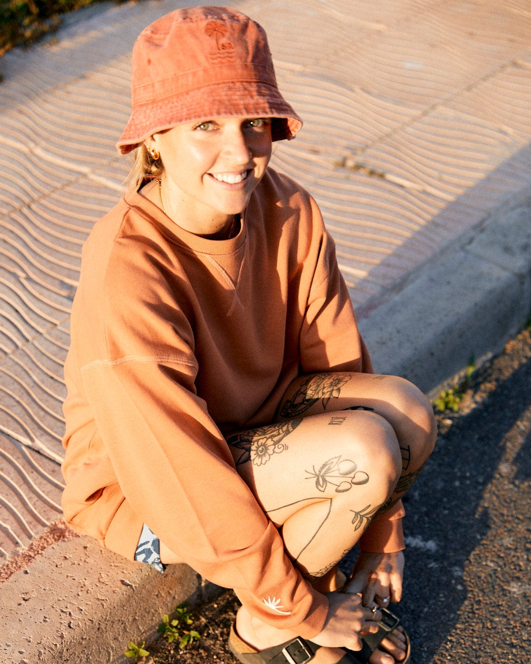 Person wearing an orange bucket hat and the Saltrock Journey - Womens Sweatshirt - Peach with embroidered graphics, squatting on the curbside with tattoos visible on their legs, and smiling at the camera. The relaxed fit adds to their effortlessly cool vibe.