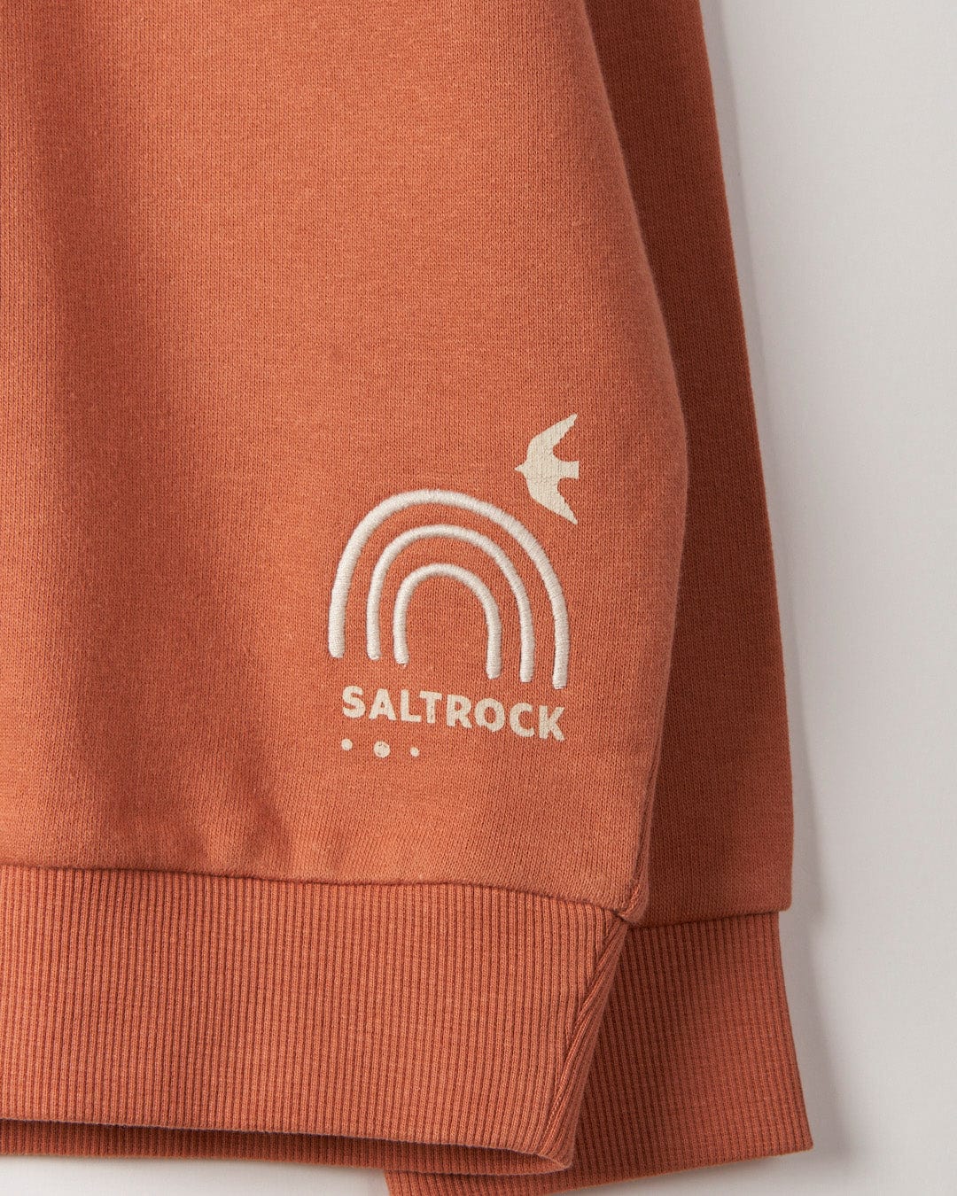 Close-up of the corner of a rust-colored, 100% cotton sweater featuring embroidered graphics with the word "Saltrock," a rainbow design, and a bird.