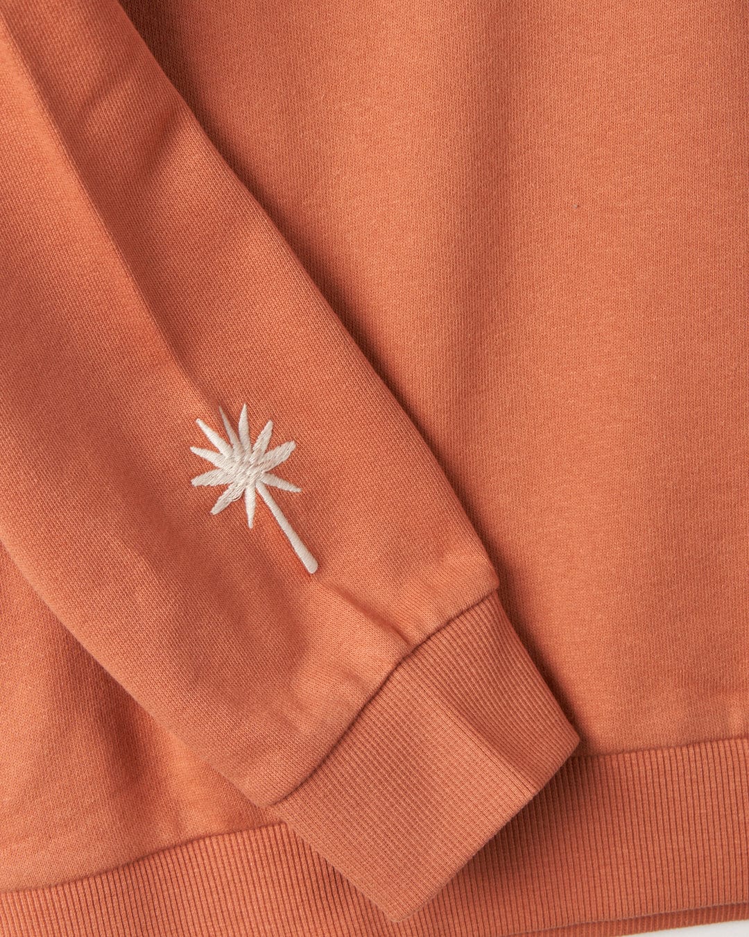 Close-up of a rust-colored long sleeve shirt with a white embroidered palm tree on the cuff. This relaxed fit Journey - Womens Sweatshirt - Peach from Saltrock, made from 100% cotton, beautifully showcases the intricate embroidered graphics for a stylish look.