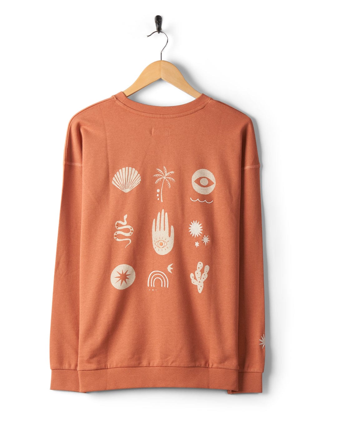 A Saltrock Journey - Womens Sweatshirt - Peach hangs on a wooden hanger, showcasing 100% cotton comfort. The back features embroidered graphic symbols in white, including a palm tree, a hand, a snake, and celestial elements. Enjoy the relaxed fit of this unique piece.