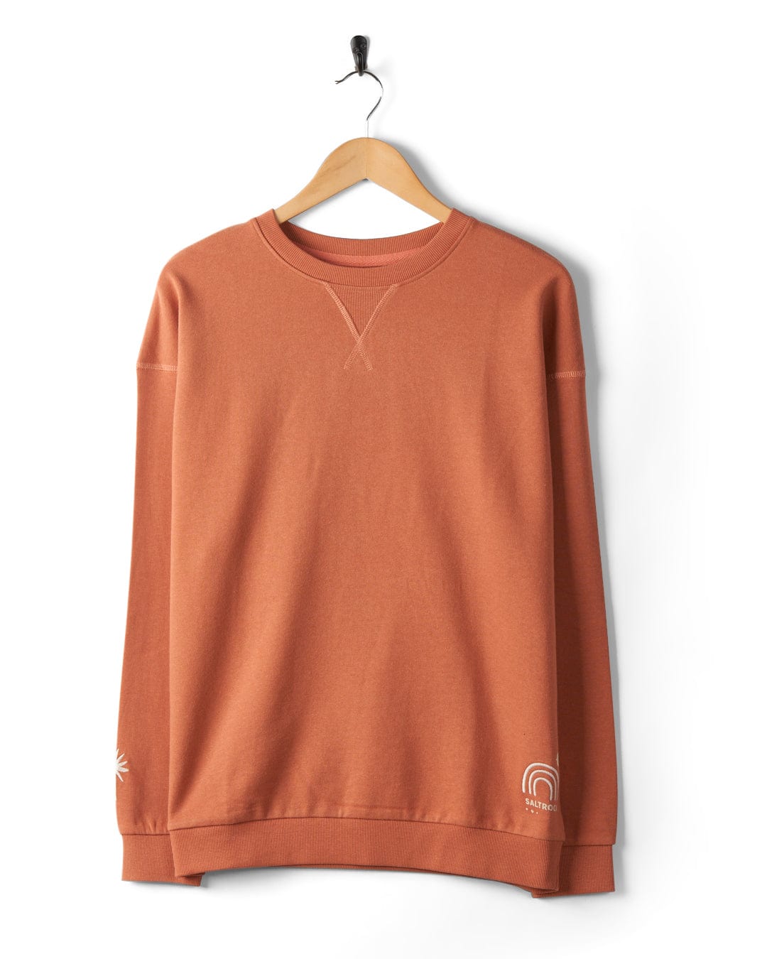A Journey - Womens Sweatshirt - Peach, made from 100% cotton with a relaxed fit, hangs on a wooden hanger against a white background. It features delicate embroidered graphics near the left wrist and bottom right hem.