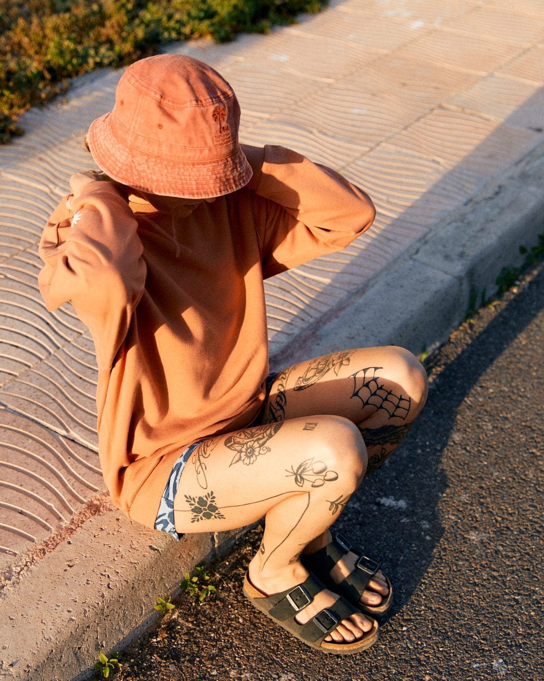Person wearing an orange bucket hat and matching sweatshirt, sitting on the curb. The relaxed fit Saltrock Journey - Womens Sweatshirt - Peach is 100% cotton and features embroidered graphics. Legs are tattooed with various designs, including a spiderweb and flowers. Wearing black sandals.
