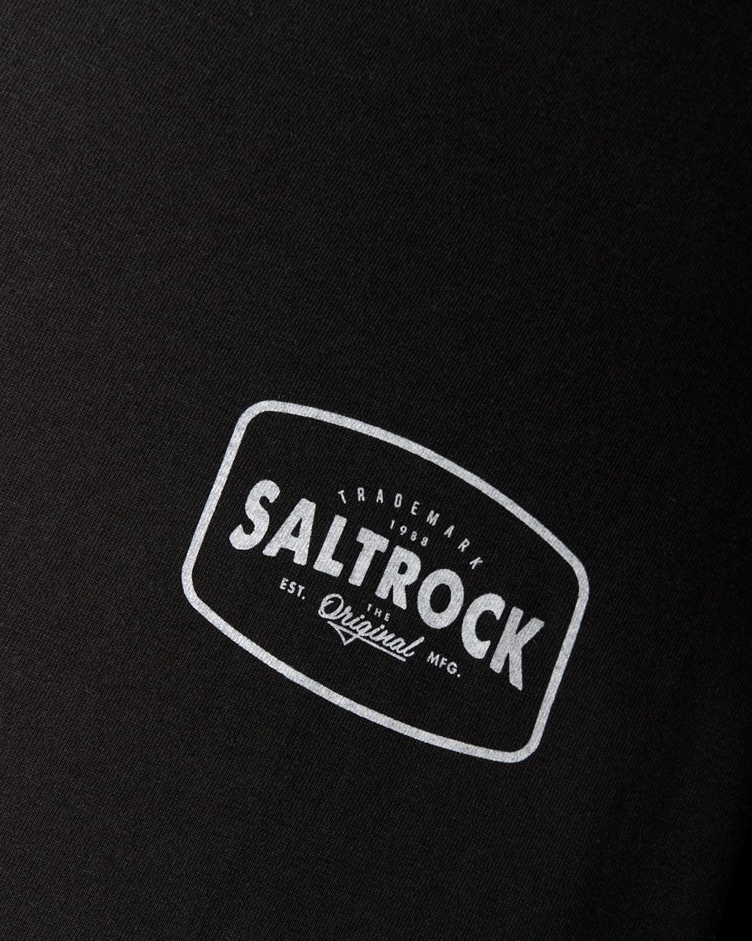 Close-up of the black fabric from the Jungle Wilds 2 Men's T-Shirt by Saltrock, featuring white branding that includes the text "Trademark Saltrock," "Original," and "Est. 1988 Mfg." inside a rounded rectangular border.