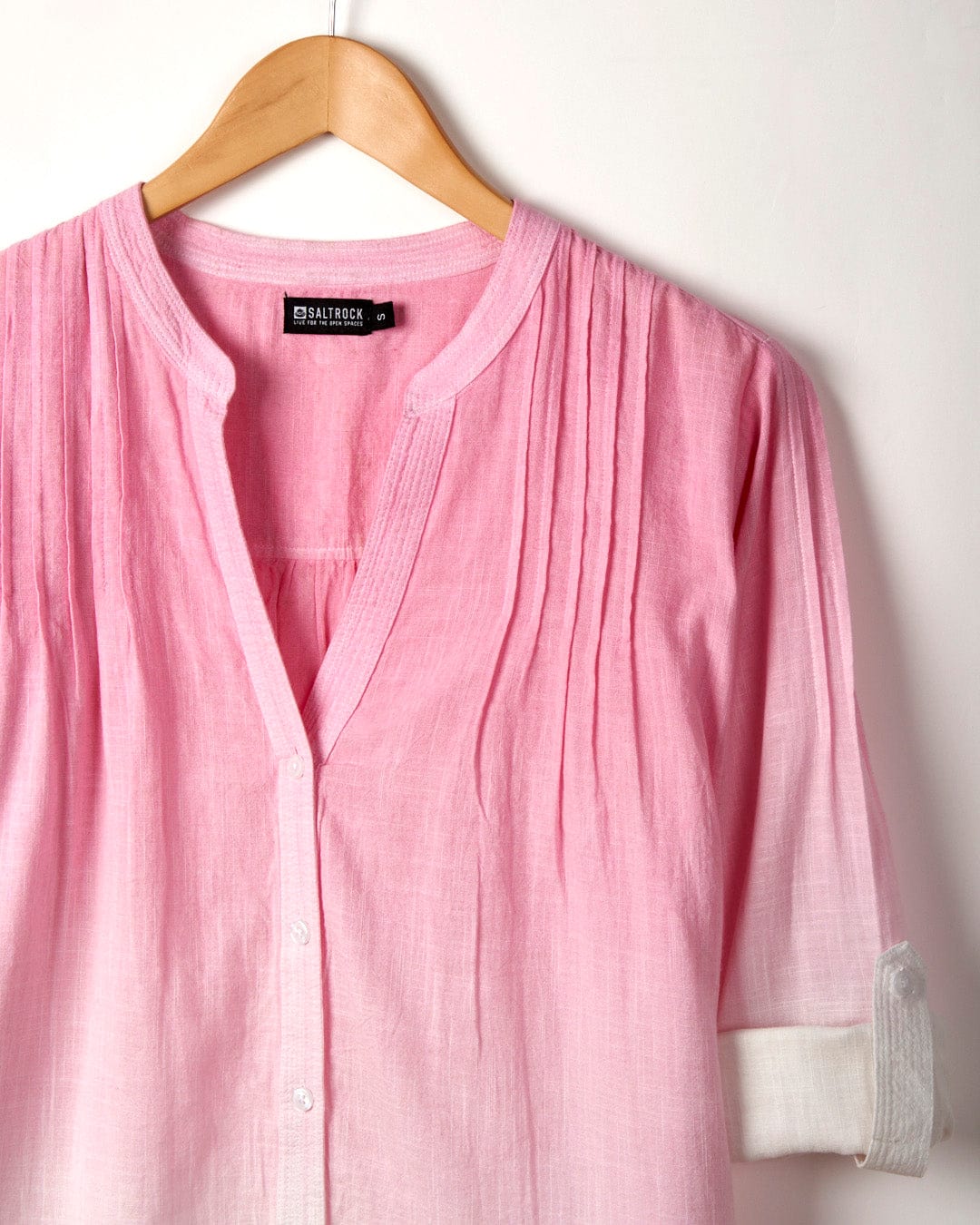 A Karthi - Womens Long Sleeve Shirt - Pink/White with vertical pleats, hanging on a wooden hanger. It features a white cuffed sleeve, an oversized fit, and a 'Saltrock' label inside the collar, made from lightweight cotton material.
