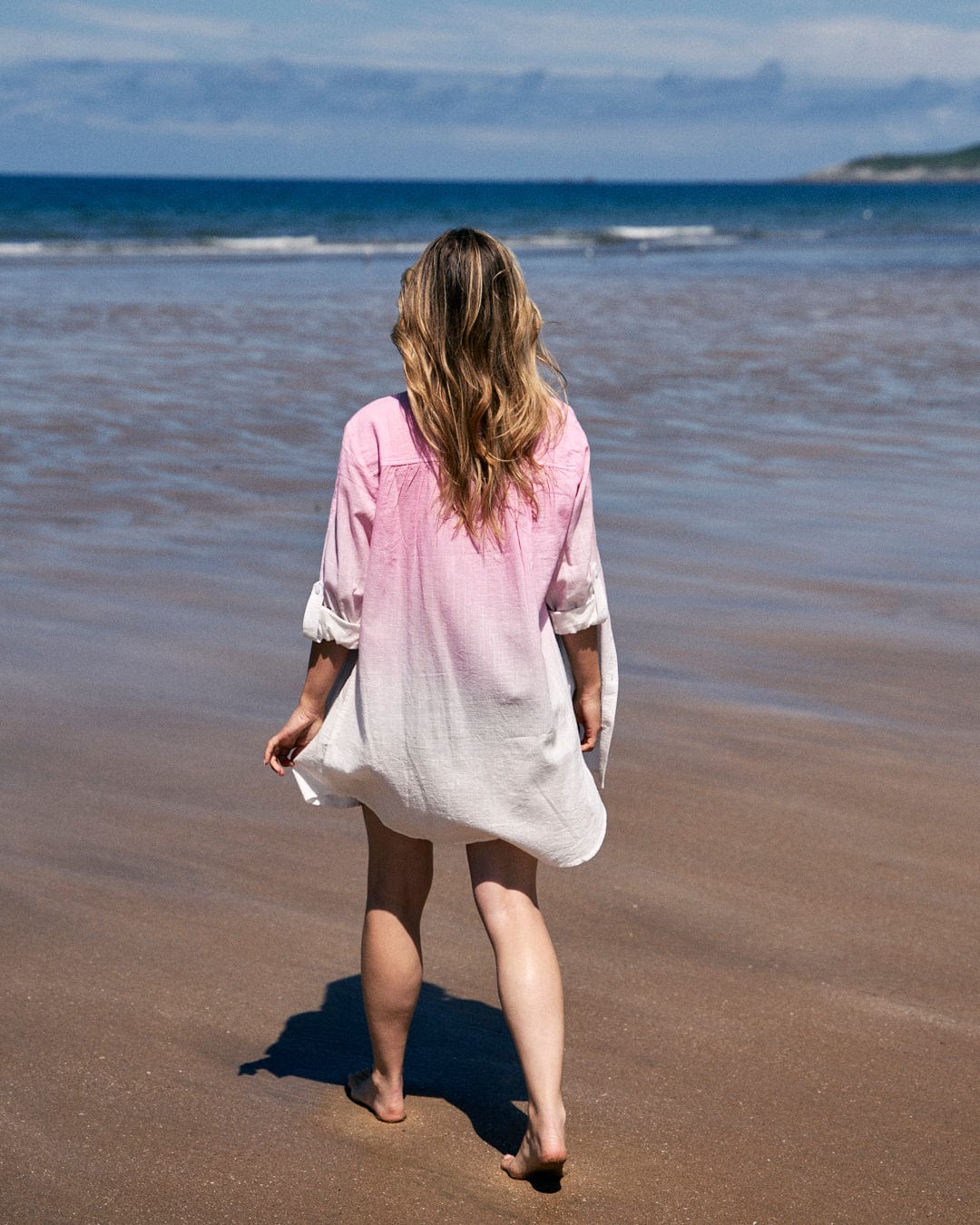A person with long hair wearing the Saltrock Karthi - Womens Long Sleeve Shirt - Pink/White made of lightweight cotton material walks barefoot on a sandy beach towards the ocean.