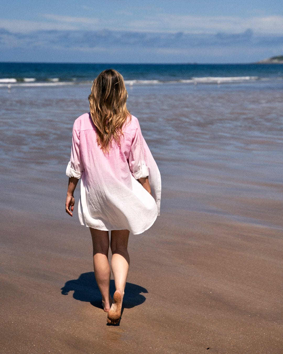 A person with long hair wearing a Saltrock Karthi - Womens Long Sleeve Shirt - Pink/White made of lightweight cotton material walks barefoot along a sandy beach towards the ocean.