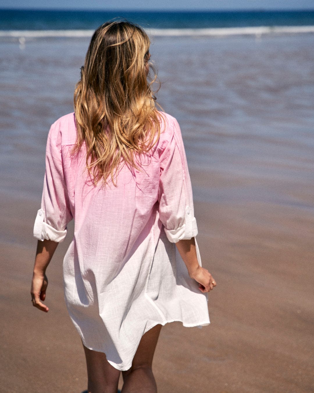 A woman with long hair walks on a beach, wearing a Saltrock Karthi - Womens Long Sleeve Shirt - Pink/White.