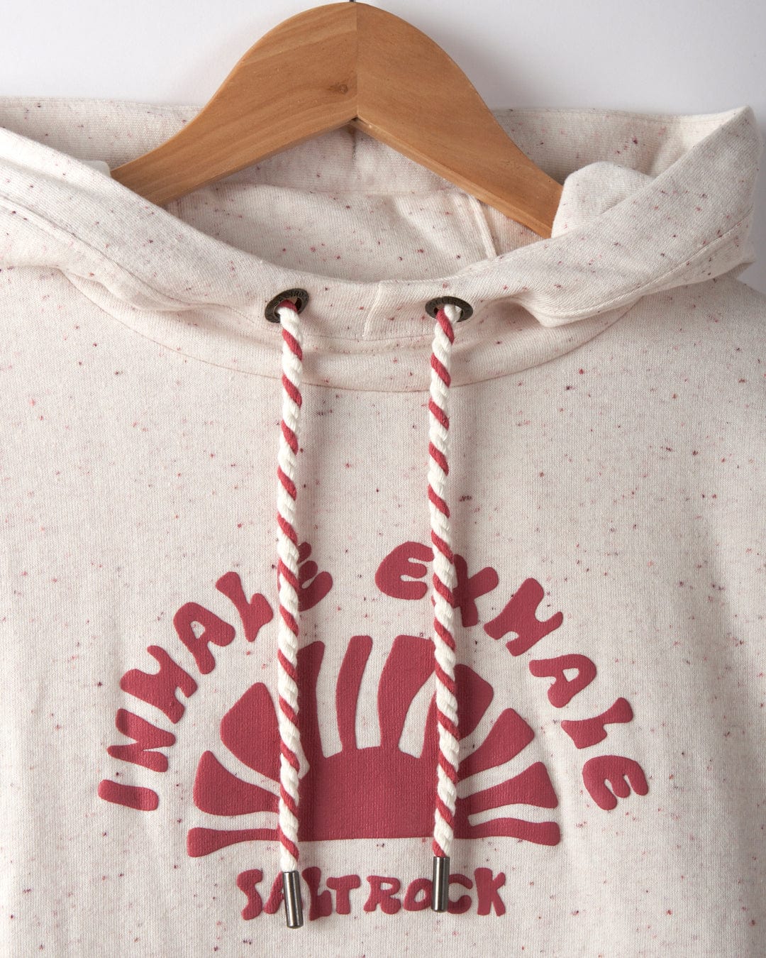 Close-up of the Saltrock Kellie Women's Cropped Pop Hoodie in Cream, showcasing a speckled design with "INHALE EXHALE" in Saltrock puffed print and a red graphic on the front. The hoodie features striped drawstrings, is cropped at the waist, and is hanging on a wooden hanger.