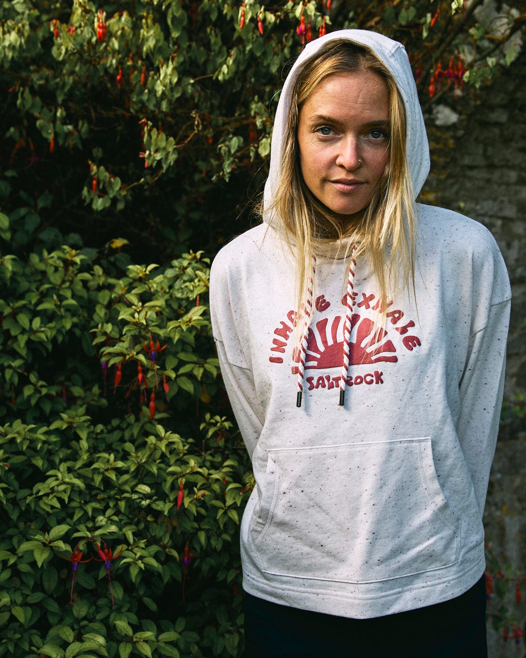 A woman with blonde hair stands in front of a leafy backdrop, wearing the Kellie Hoodie from Saltrock, a cream-colored cropped hoodie featuring red text and a Saltrock puffed print. Made from recycled materials, she has her hands in her pockets and looks directly at the camera.